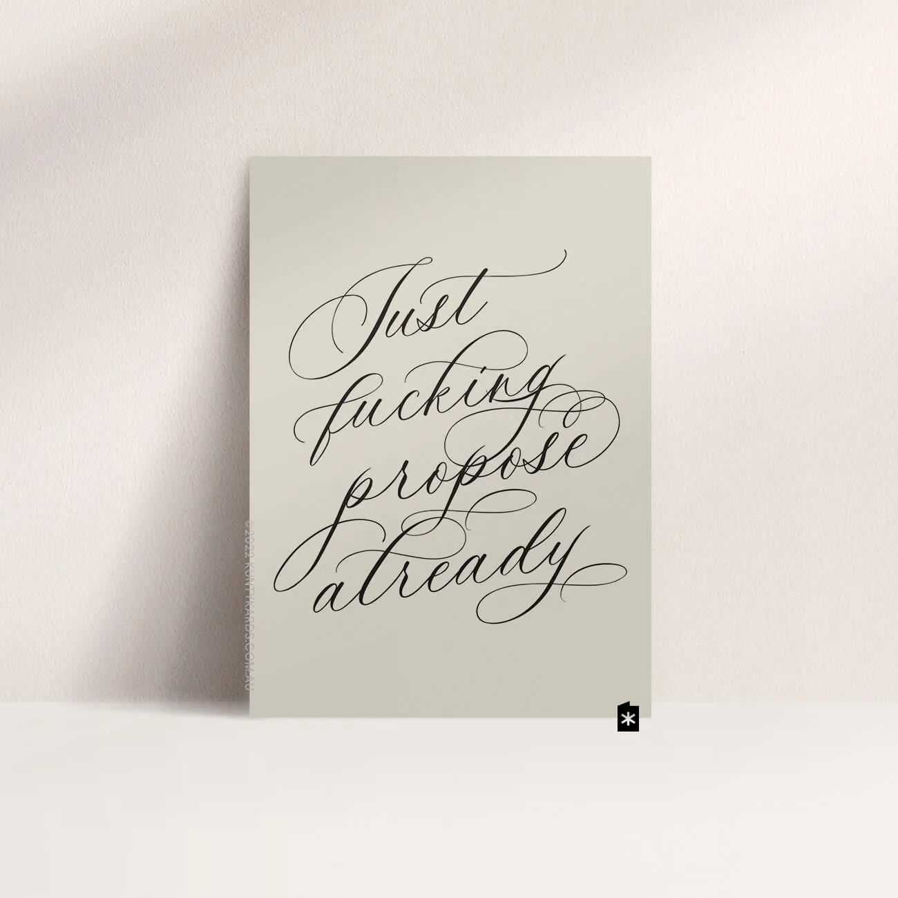 Grey card with black ink printed in script font on the front that says "Just fucking propose already" made in Perth WA by Kunty Kards
